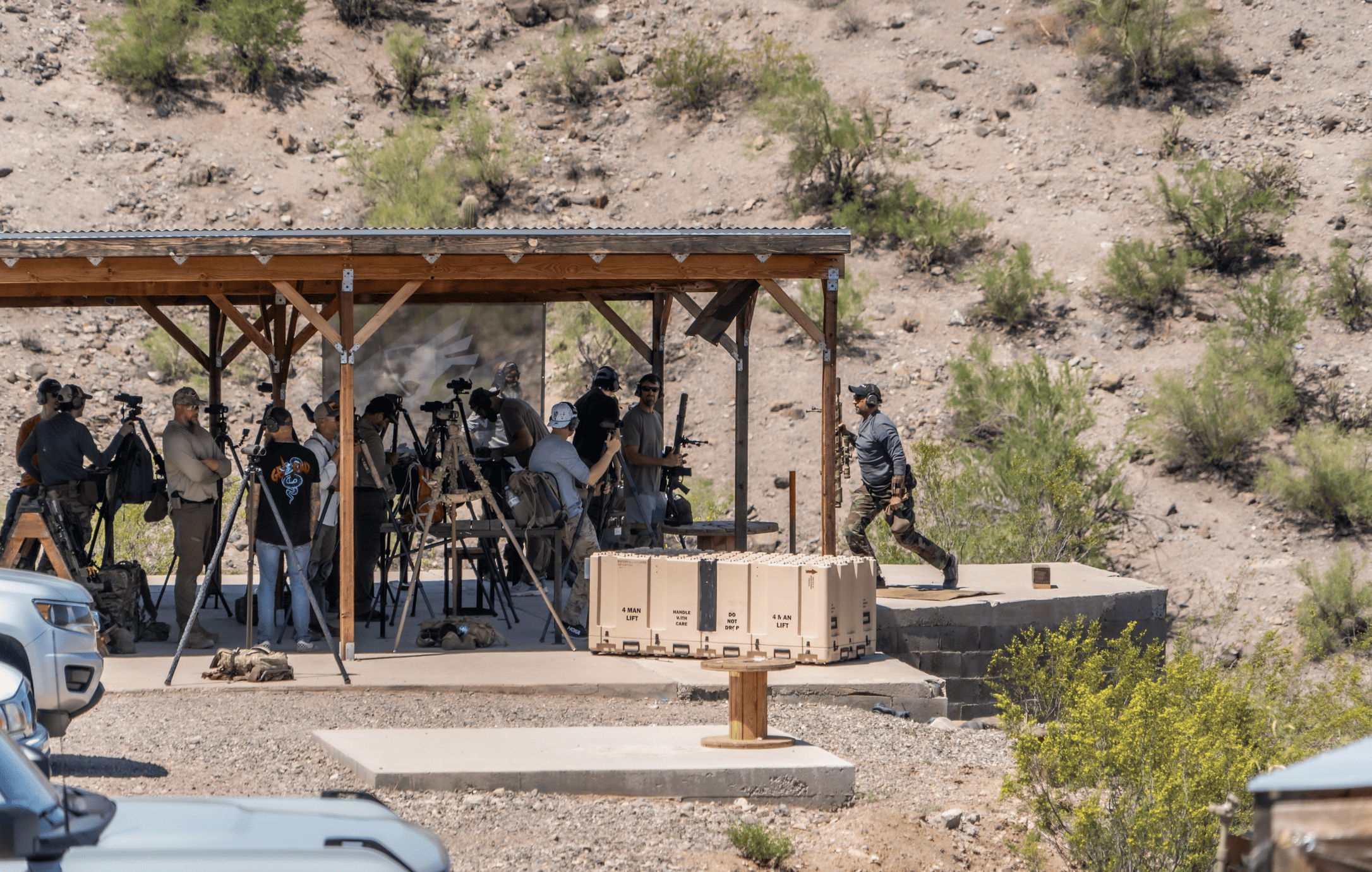 ad3ea8dd-efce-4e6d-a0d1-fbca794dc804.webp A group of shooters and instructors gather with tripods, spotting scopes, and gear under a rustic wooden shade structure at an outdoor desert training bay.