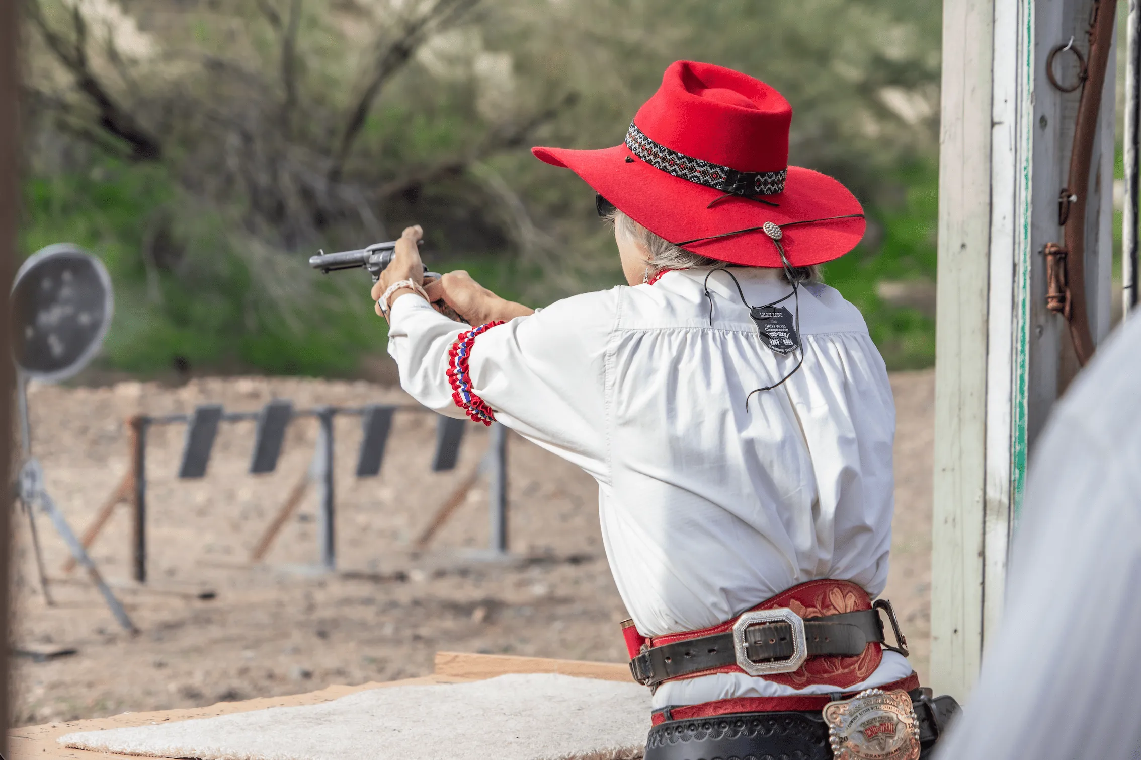 Competitor in red cowboy hat aiming a single-action revolver downrange at targets during a cowboy action match.