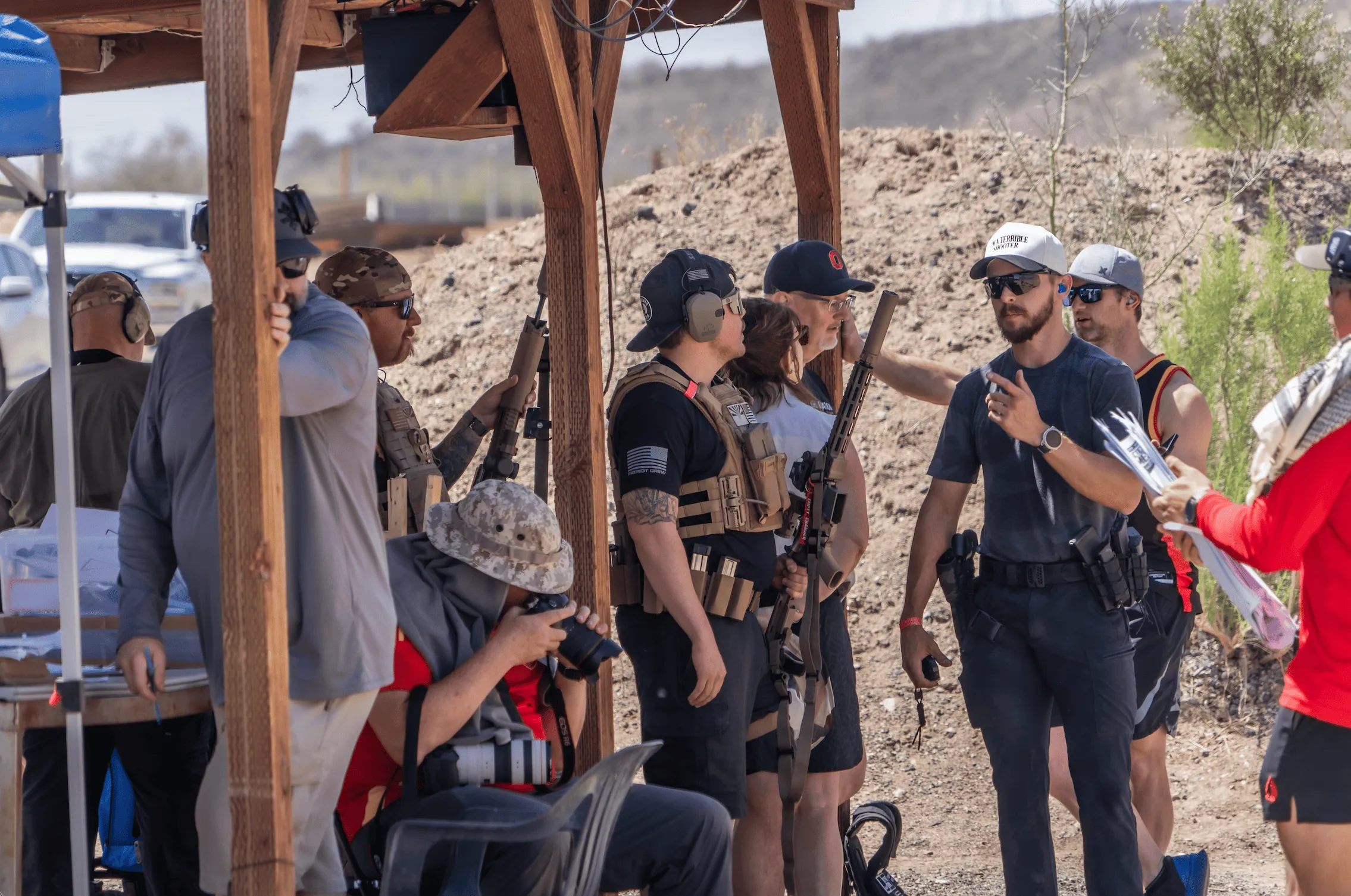 A community of shooters and instructors gathered under a wooden shade canopy in a high-desert bay, conversing and staging their slung rifle platforms and tactical gear before a training run.