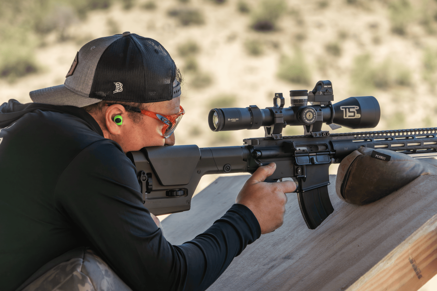 Close-up of a shooter with eye and ear protection aiming a scoped rifle system stabilized on a support bag from a wooden shooting platform.