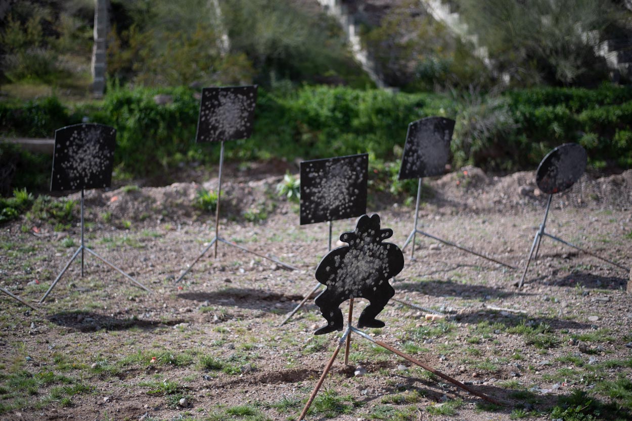 An outdoor shooting range features several black metal targets scattered across a dirt and sparse grass field. In the foreground, a prominent target is shaped like a stylized cartoon silhouette of a person wearing a wide-brimmed hat. Behind it, five square, rectangular, and round metal plates are positioned at varying distances, all mounted on metal tripods. Every target is heavily pockmarked with white splatter marks from repeated bullet impacts. The background consists of a sloping hill with green brush and a concrete barrier structure under soft, diffused daylight.
