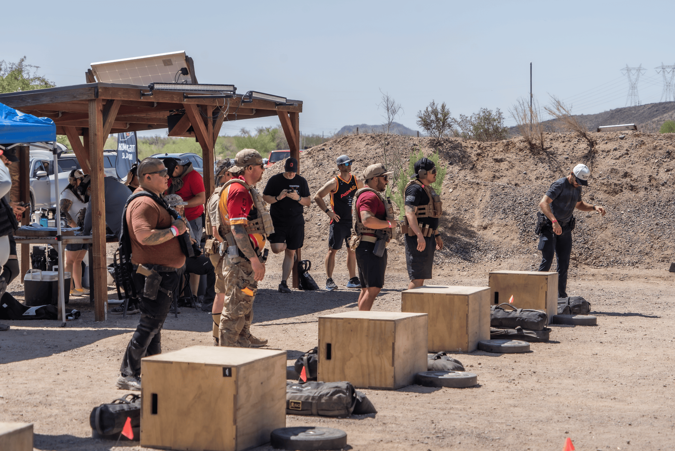 A lineup of competitors wearing tactical gear, plate carriers, and baseball caps standing on a hot dirt deck during an outdoor tactical fitness and shooting competition. Wooden plyo boxes, weights, and heavy sandbags are lined up on the range under a clear sky.