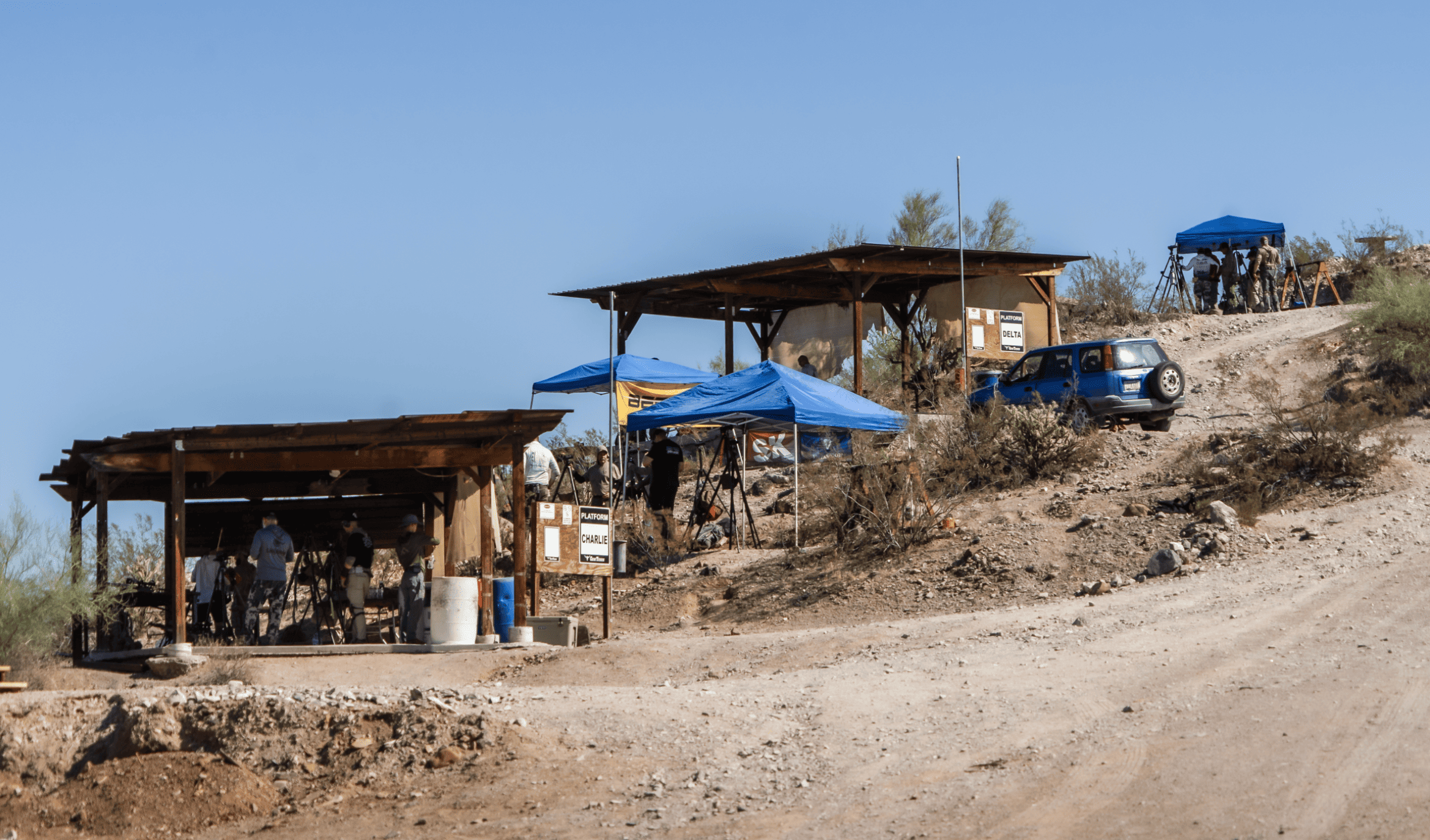 Outdoor long-range precision platforms Charlie and Delta at Cowtown Range, featuring rustic wooden shade structures, blue pop-up tents, a parked SUV, and shooters preparing gear on a rugged high-desert hillside.