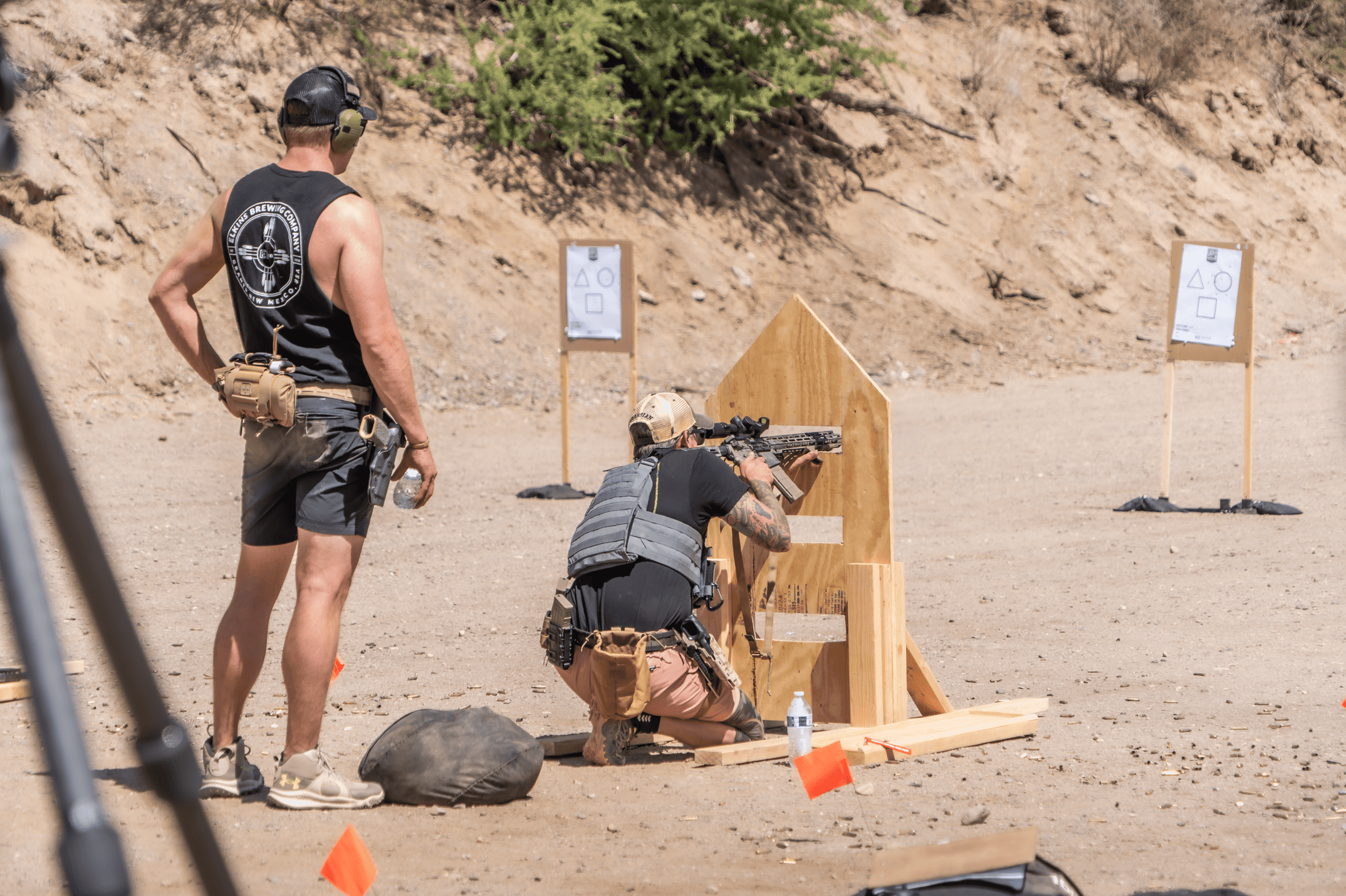 A rifleman kneeling in a gray plate carrier and camo cap fires through a custom wooden step-barricade, while a spotter in a black graphic tank top stands by to track target impacts at an outdoor tactical training facility.