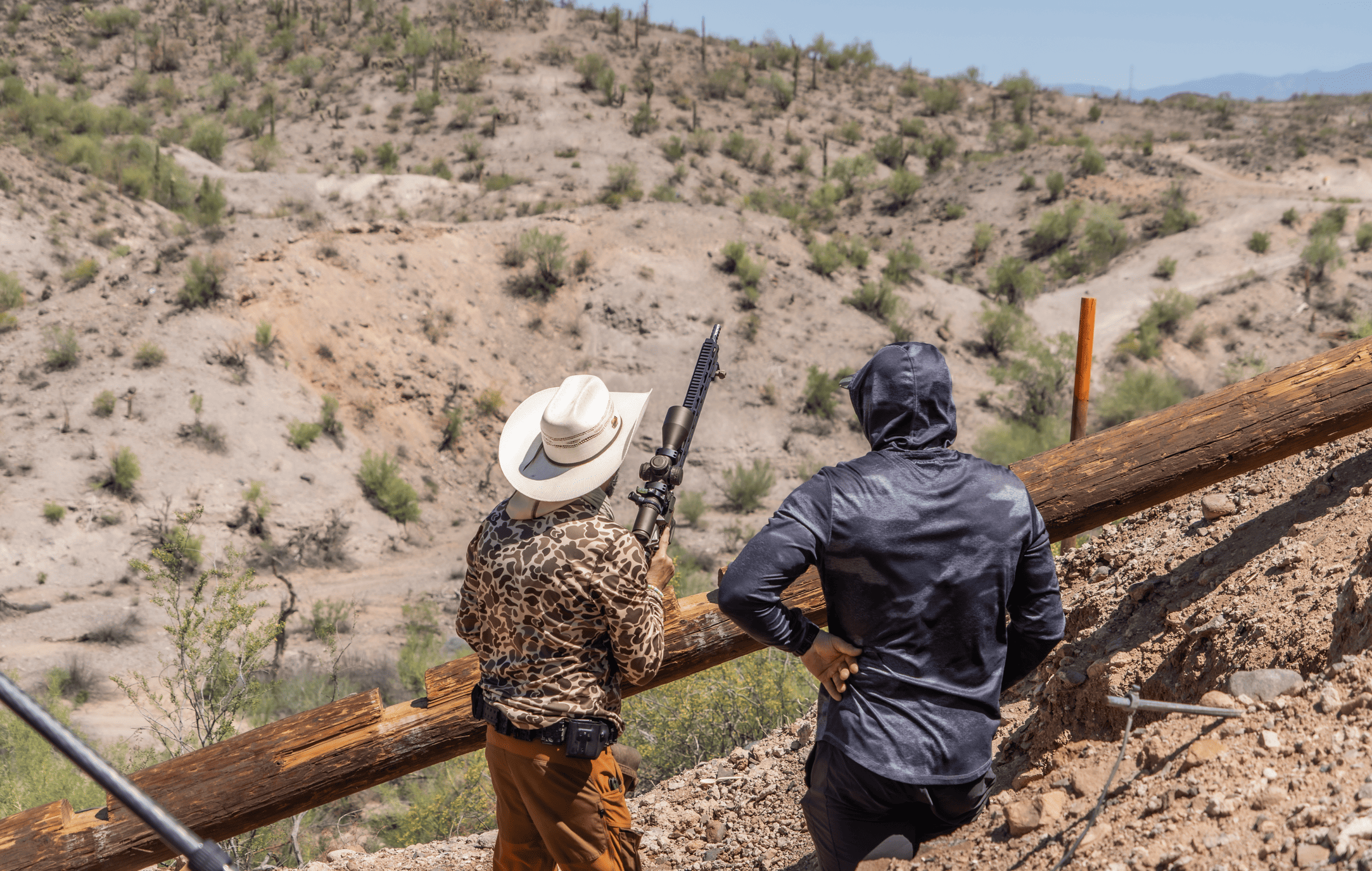 Two shooters tracking targets across a vast desert canyon from a high dirt ridge. One shooter in a white cowboy hat and camo shirt readies a precision long-range rifle over a heavy wooden log barricade, while a spotter in a dark gray sun hoodie stands alongside him calling hits.