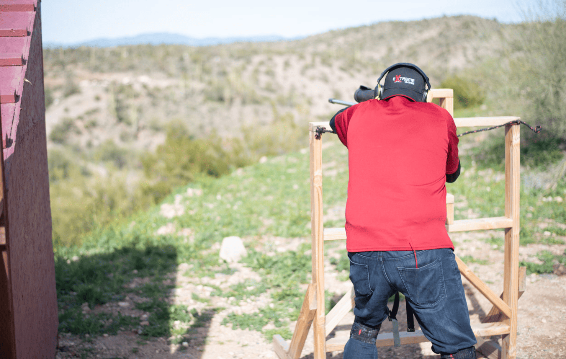 Rear view of a shooter wearing a red shirt, blue jeans, and ear protection, stabilizing a precision rifle platform on a wooden barricade over a long-range high-desert valley at Cowtown Range.