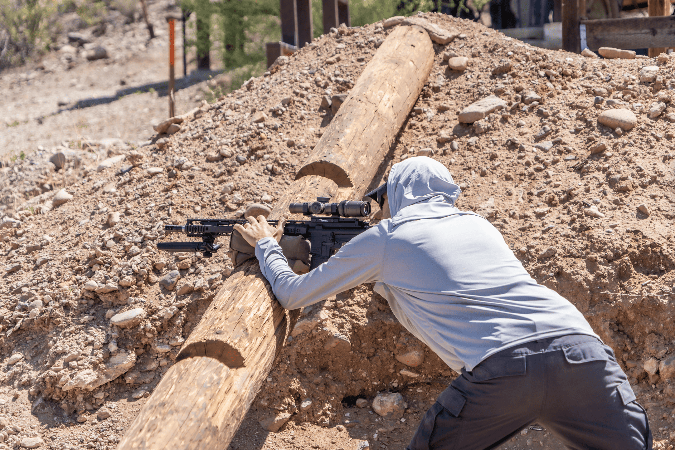 A shooter in a grey sun hoodie utilizing a sloped log barrier on a dirt mound to stabilize an AR-15 platform rifle system during an outdoor training drill.