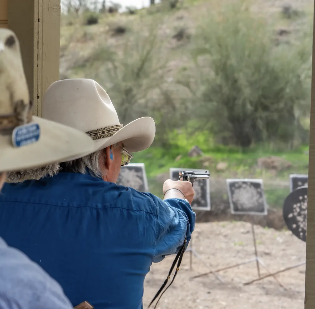 Over-the-shoulder view of a shooter in a cowboy hat aiming a single-action revolver at steel targets on a bay.