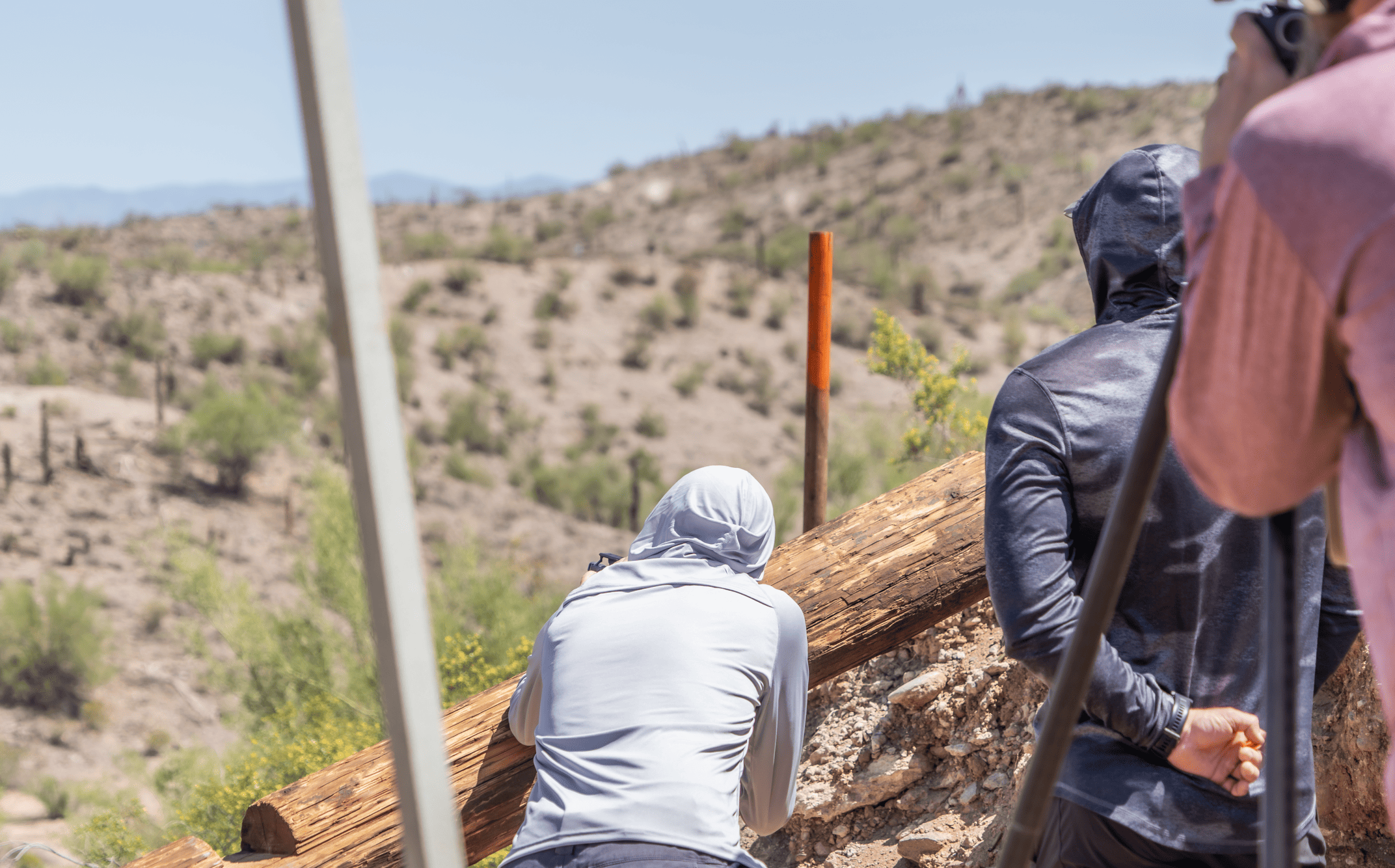 An operator in a light grey sun hoodie stabilizes a rifle system on a rustic log barricade while a spotter looks on at an outdoor desert shooting bay.