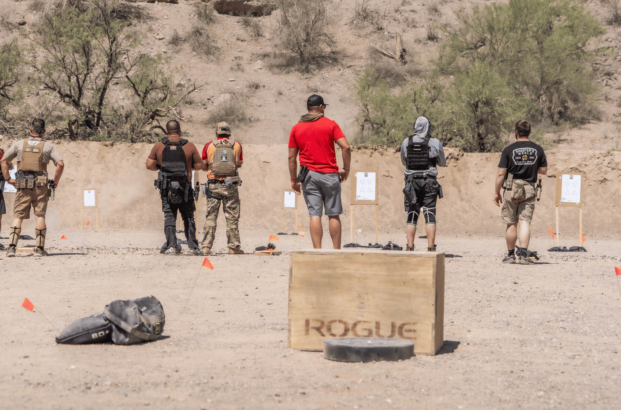 fea00785-7d69-433b-9383-a44249f797b5.webp A crew of shooters in tactical vests and everyday gear standing on the firing line, seen from behind as they prepare to engage targets downrange in a dusty desert canyon, with a heavy-duty wooden Rogue box and sandbags in the foreground.