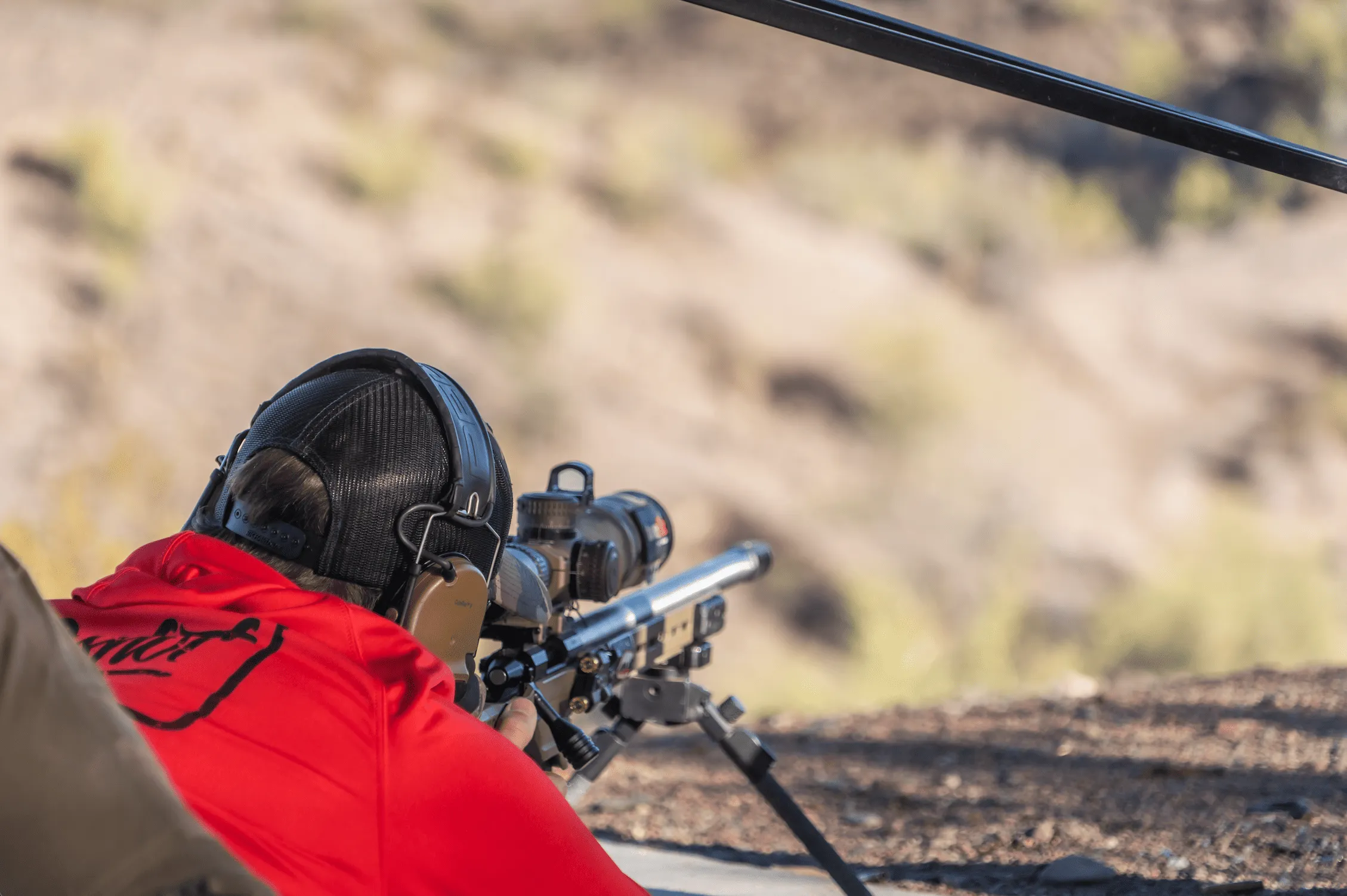 An operator wearing a red shirt and active ear protection behind a precision bolt-action rifle platform, utilizing a bipod to shoot downrange over a deep, high-desert canyon.