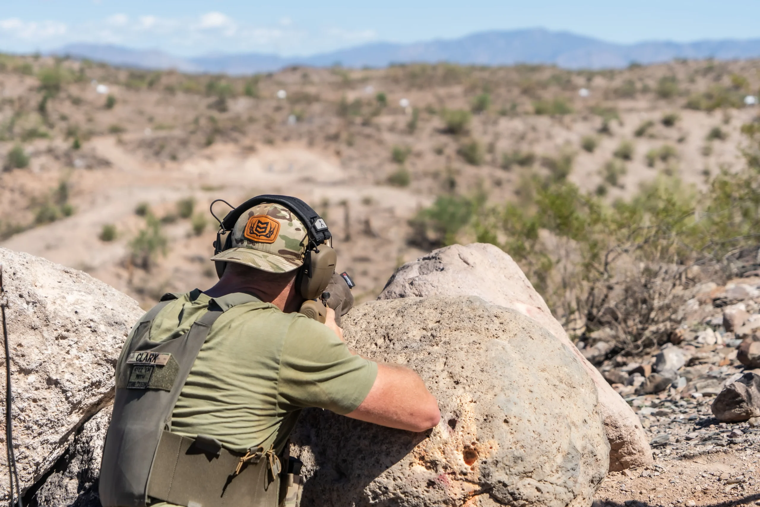 Shooter resting a precision platform on a boulder for long-range target engagement at an outdoor desert range.