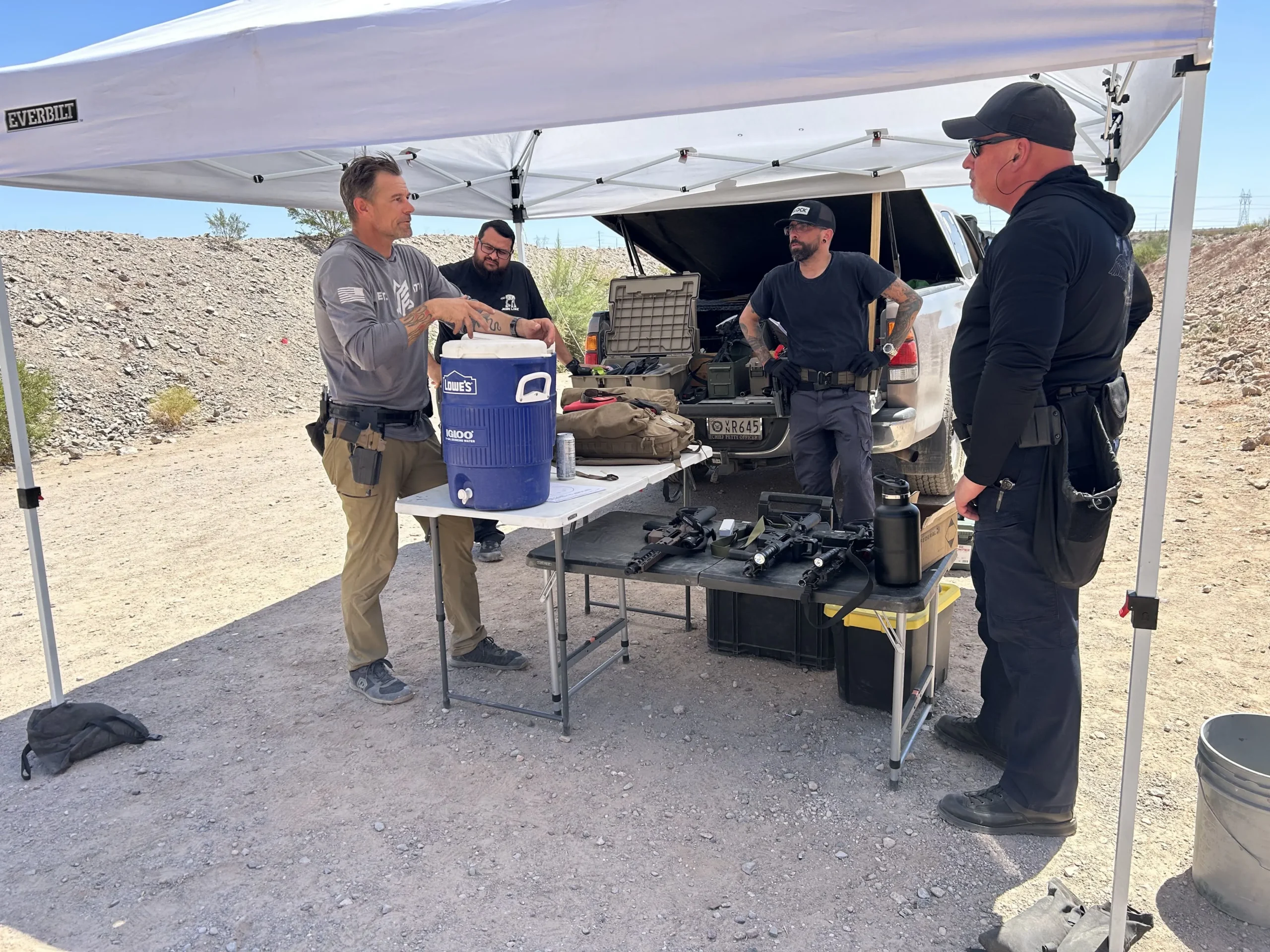 Shooters and instructors debrief under a shaded pop-up tent on an outdoor bay at Cowtown Range.