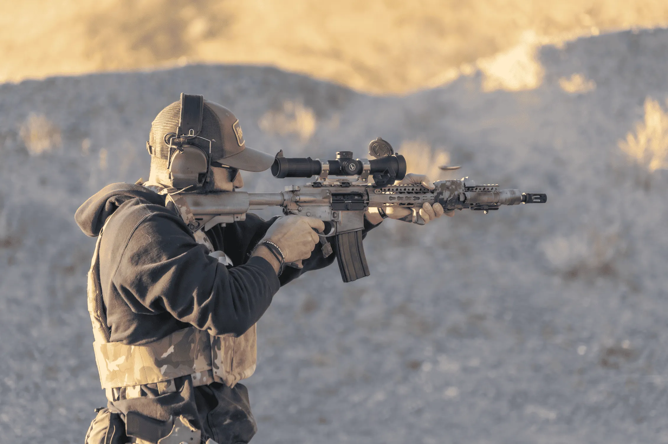 Shooter firing a scoped AR-15 carbine rifle in a high-desert bay at Cowtown Range, shell casing flying.