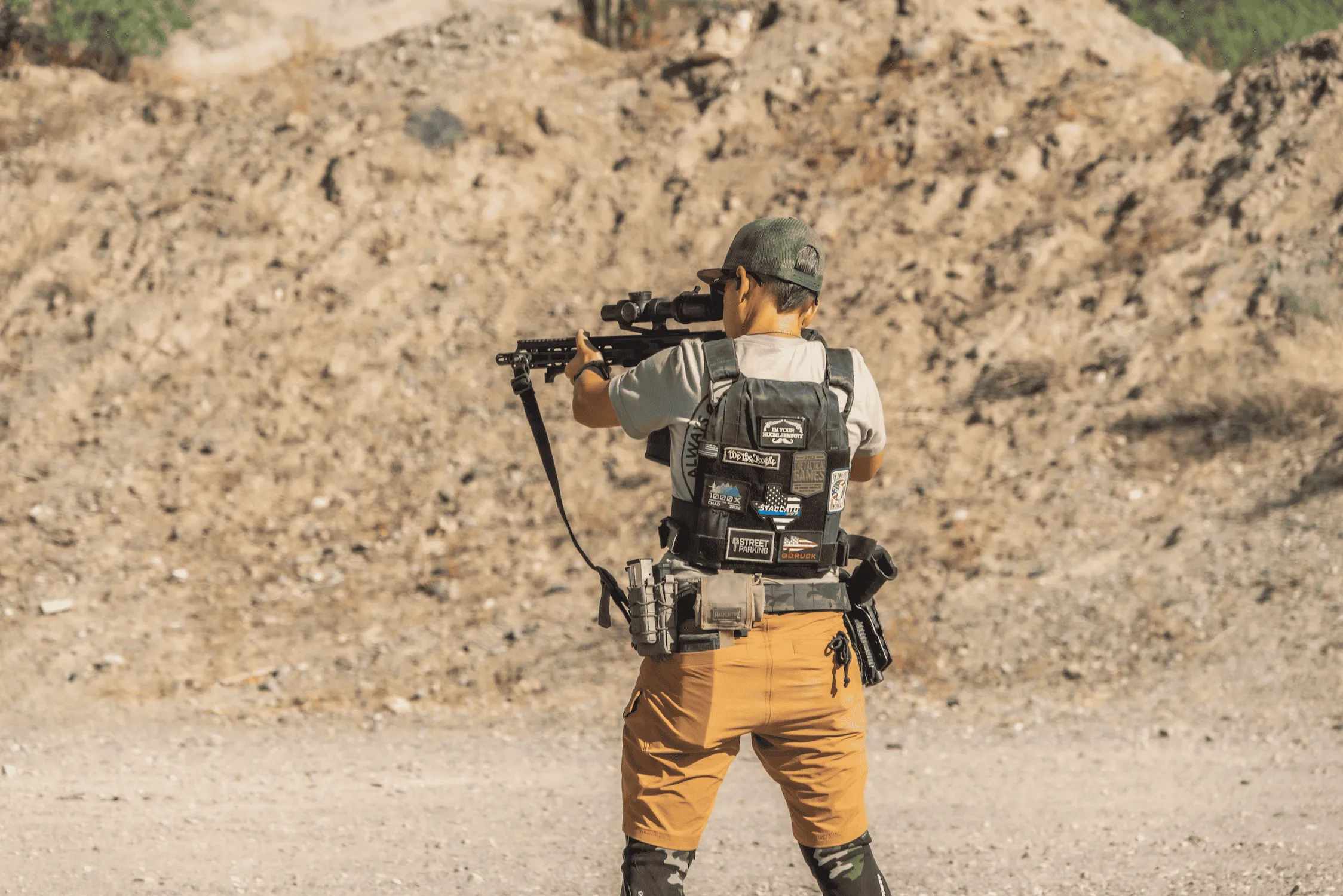 Shooter aiming a scoped rifle from the rear in a high-desert tactical outdoor bay at Cowtown Range.