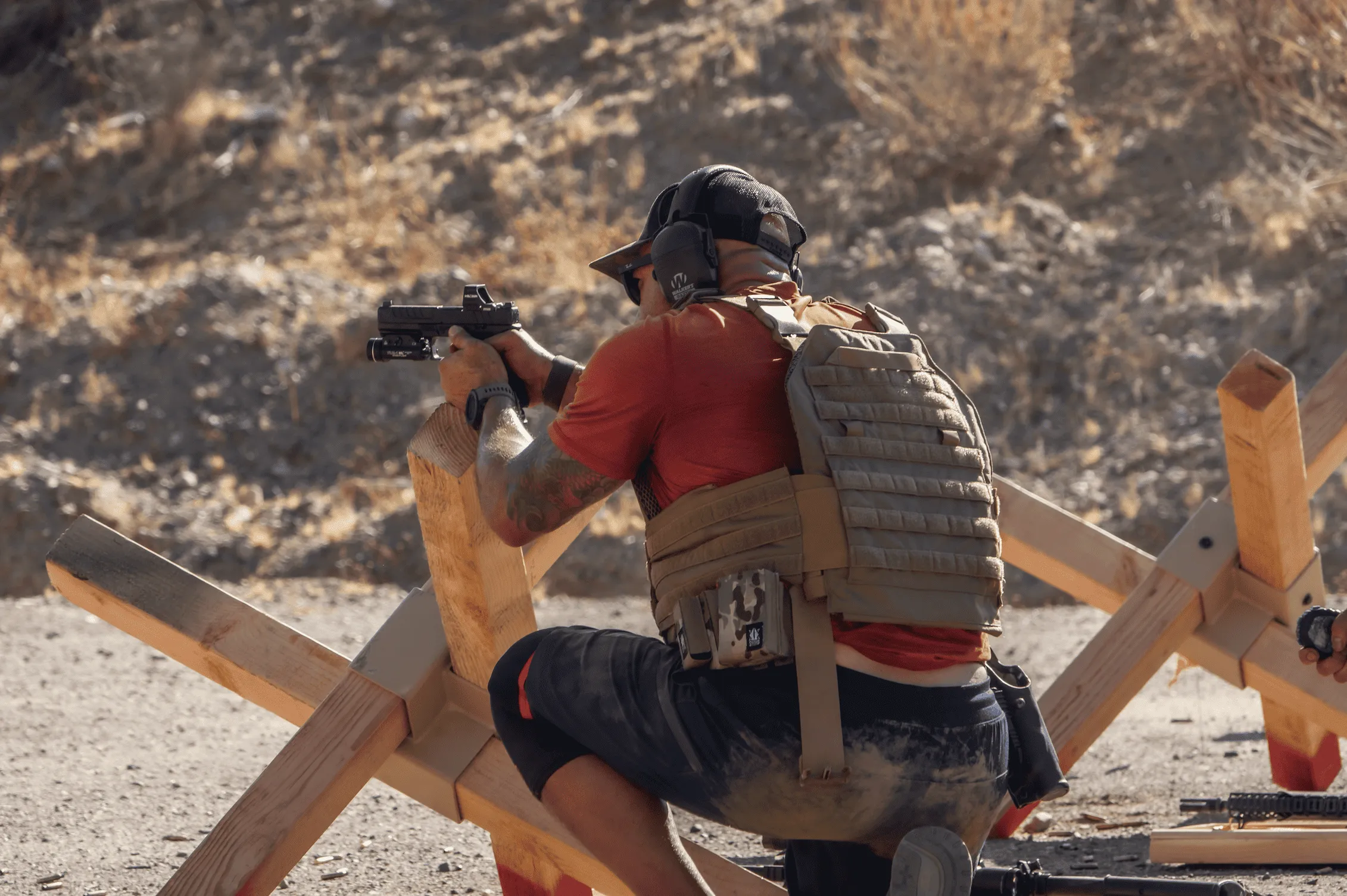 A shooter wearing a plate carrier and ear protection aims a handgun over a wooden barricade on a high-desert range.