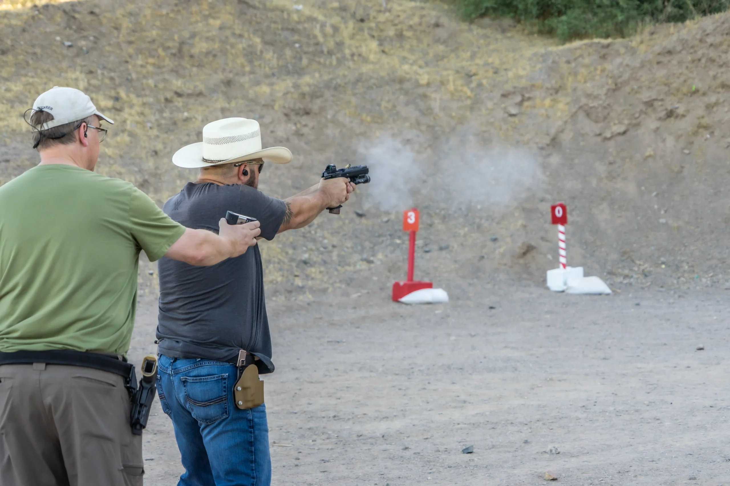 Shooter in a cowboy hat firing a red-dot handgun alongside an instructor on an outdoor range bay.