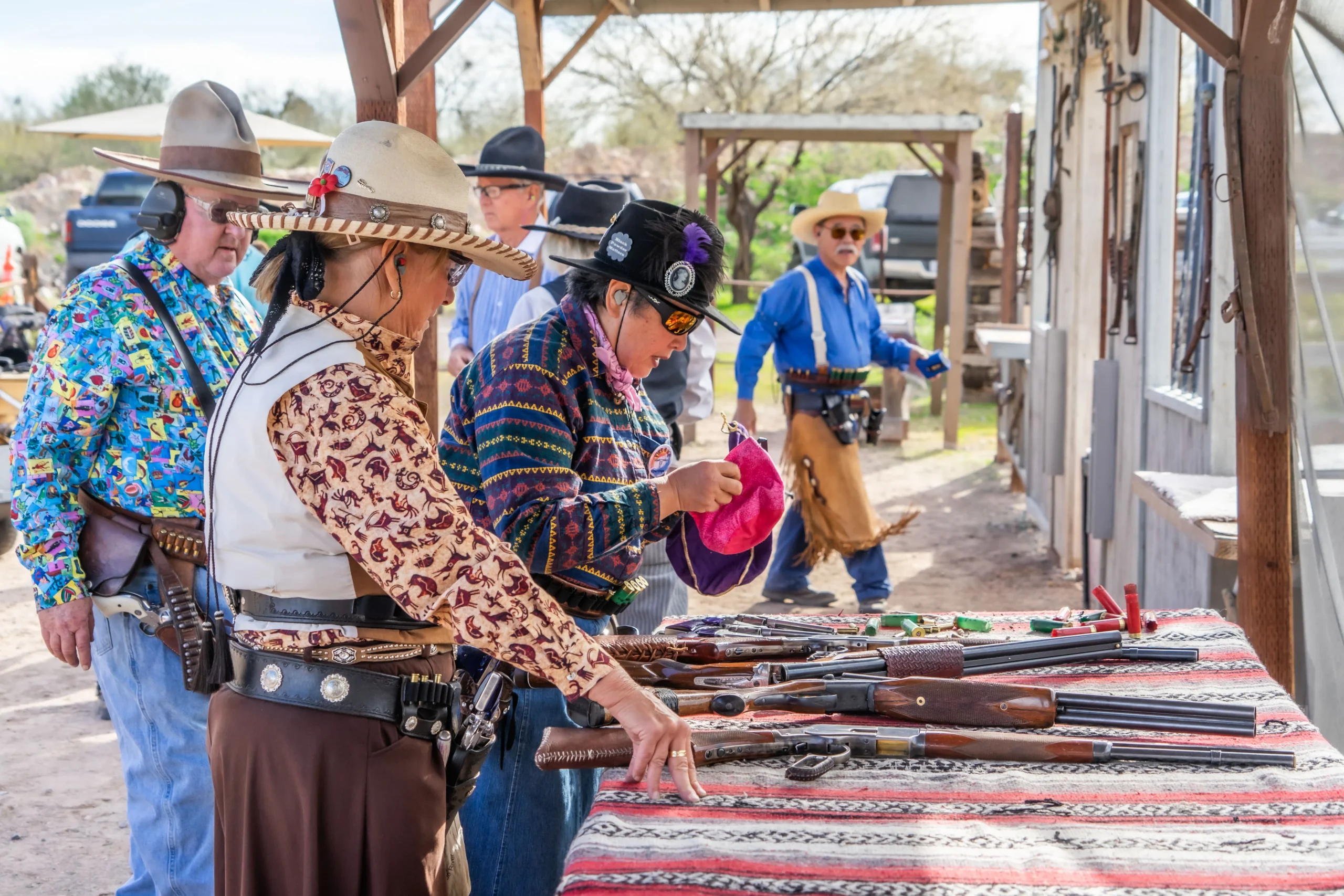 Shooters in Western attire inspecting double-barrel shotguns and lever-action rifles on a range table.