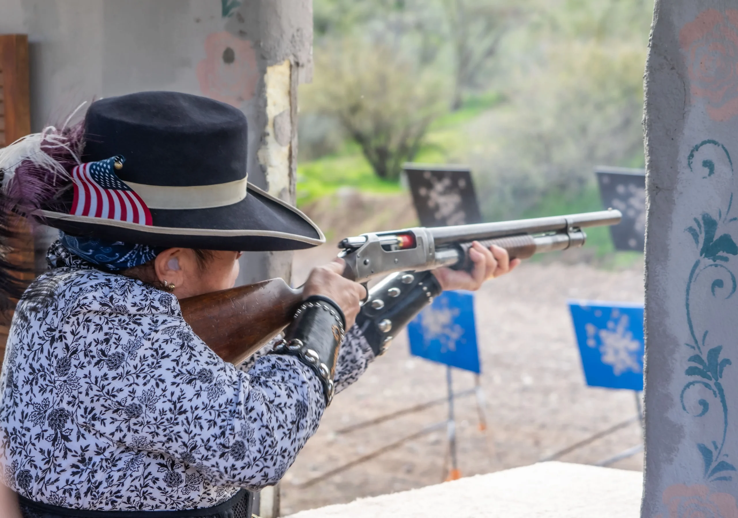 A competitor aiming a pump-action shotgun from a stage window at steel targets downrange.
