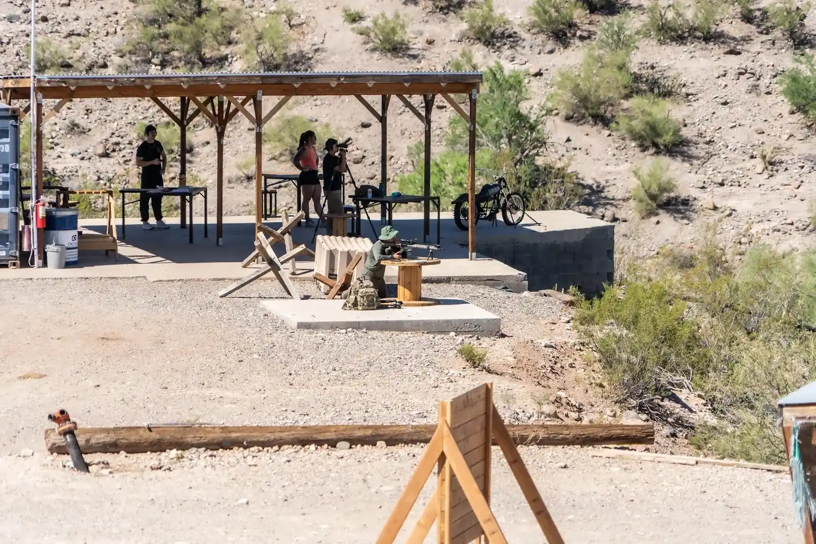 A shooter with a rifle aiming from a wooden spool bench under a covered outdoor range platform.