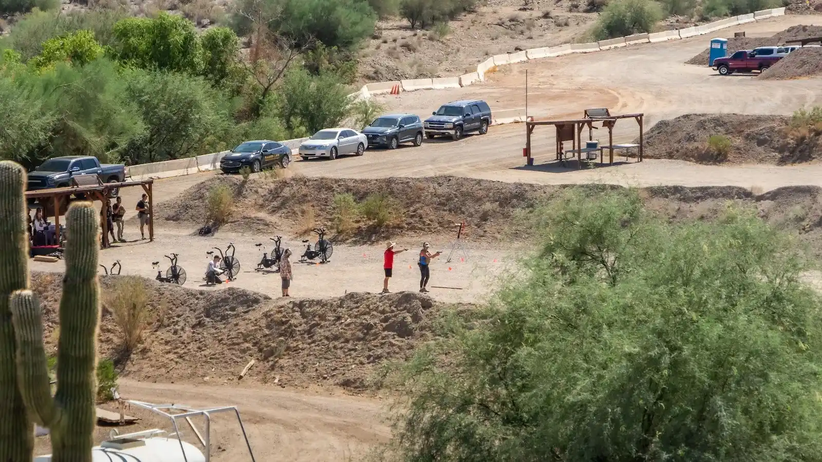 Shooters in action at a dirt outdoor shooting bay with vehicles and a Sonoran Desert hillside background.